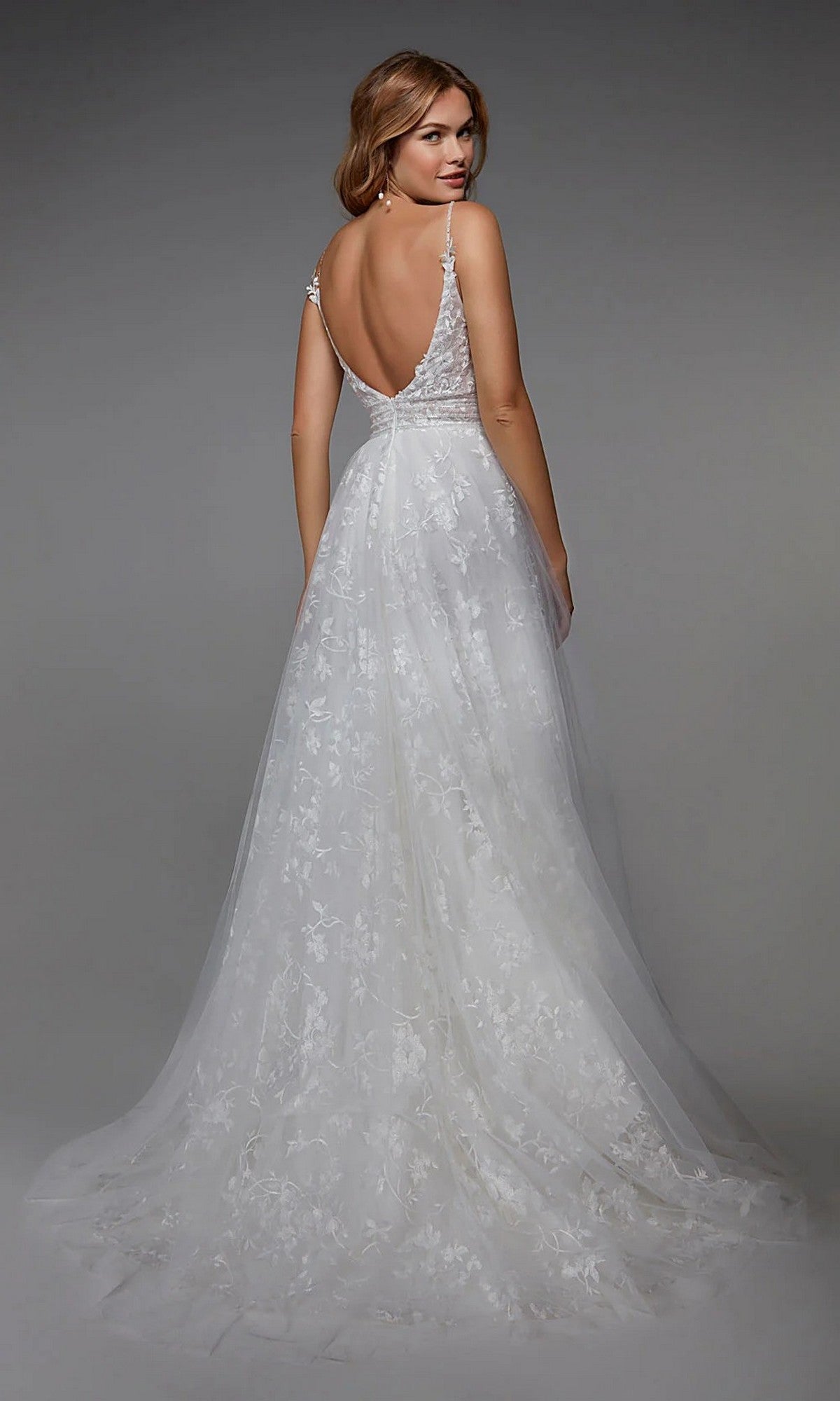 A woman stands facing away against a neutral background, wearing the Alyce 7036 Long White Dress with a sleeveless design, low back, lace detailing, and delicate floral appliqués.